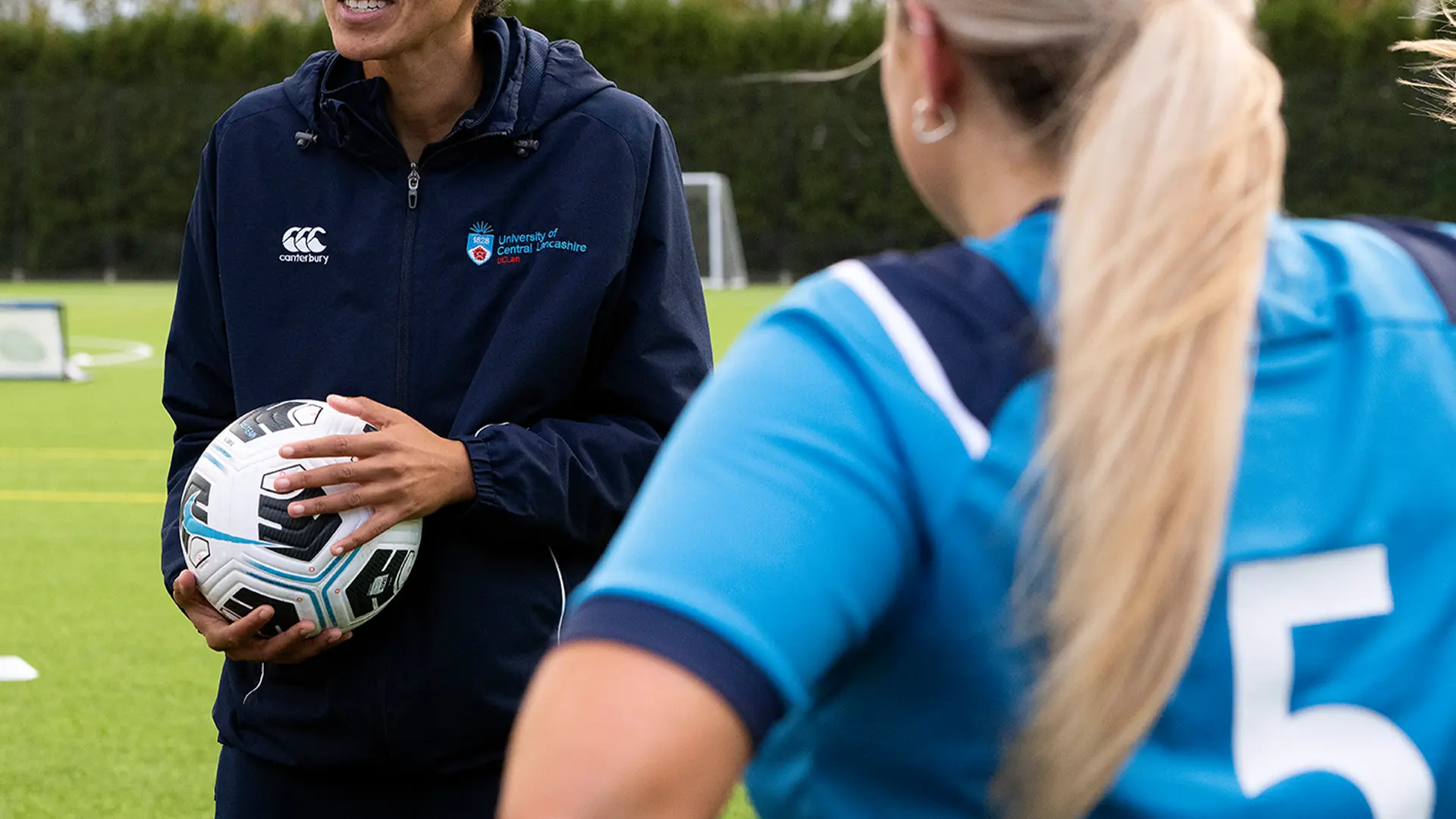 Sports coach holding football and talking to sports students on football pitch