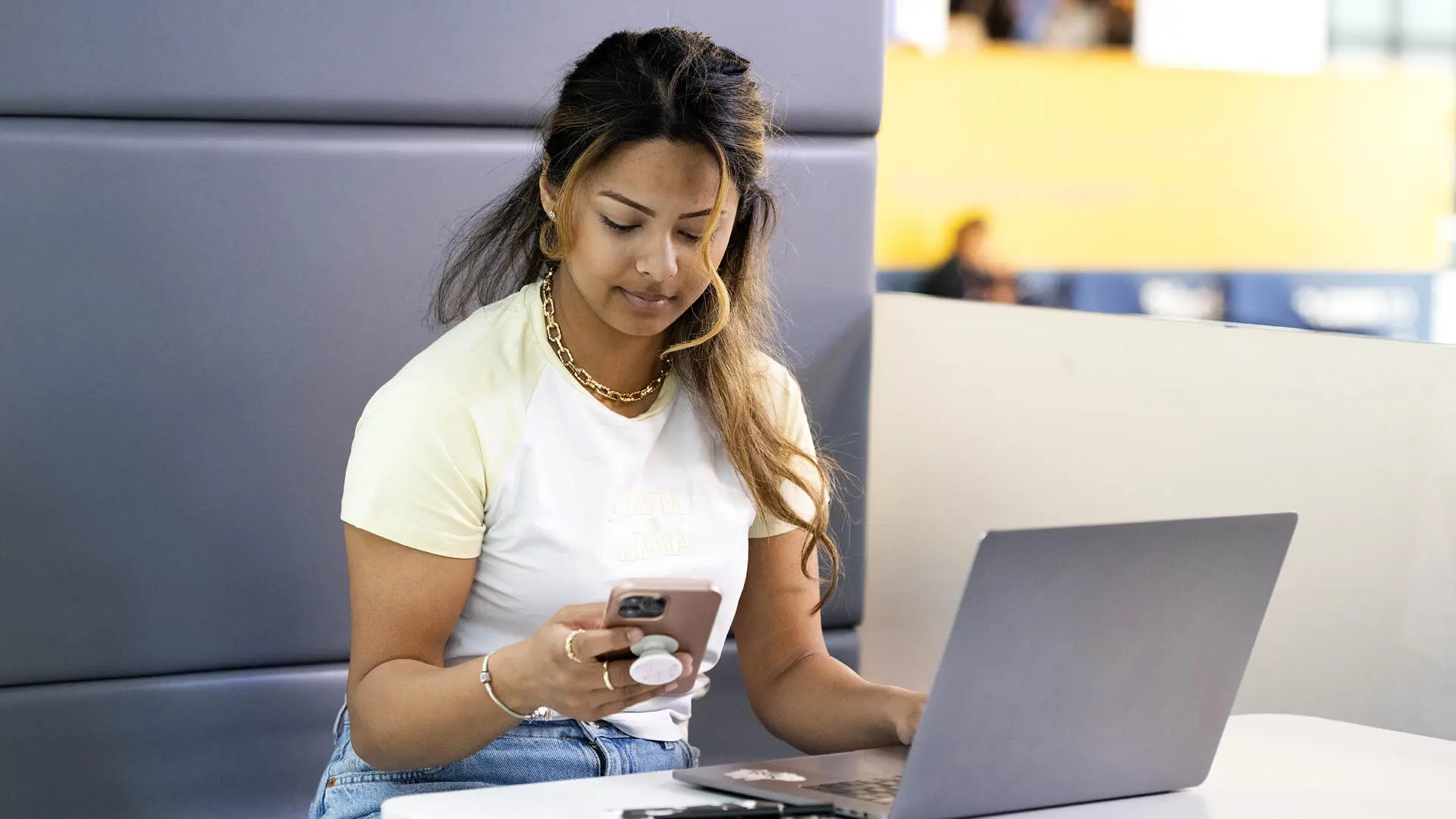 a student with long hair using a mobile phone and laptop in a social area on campus