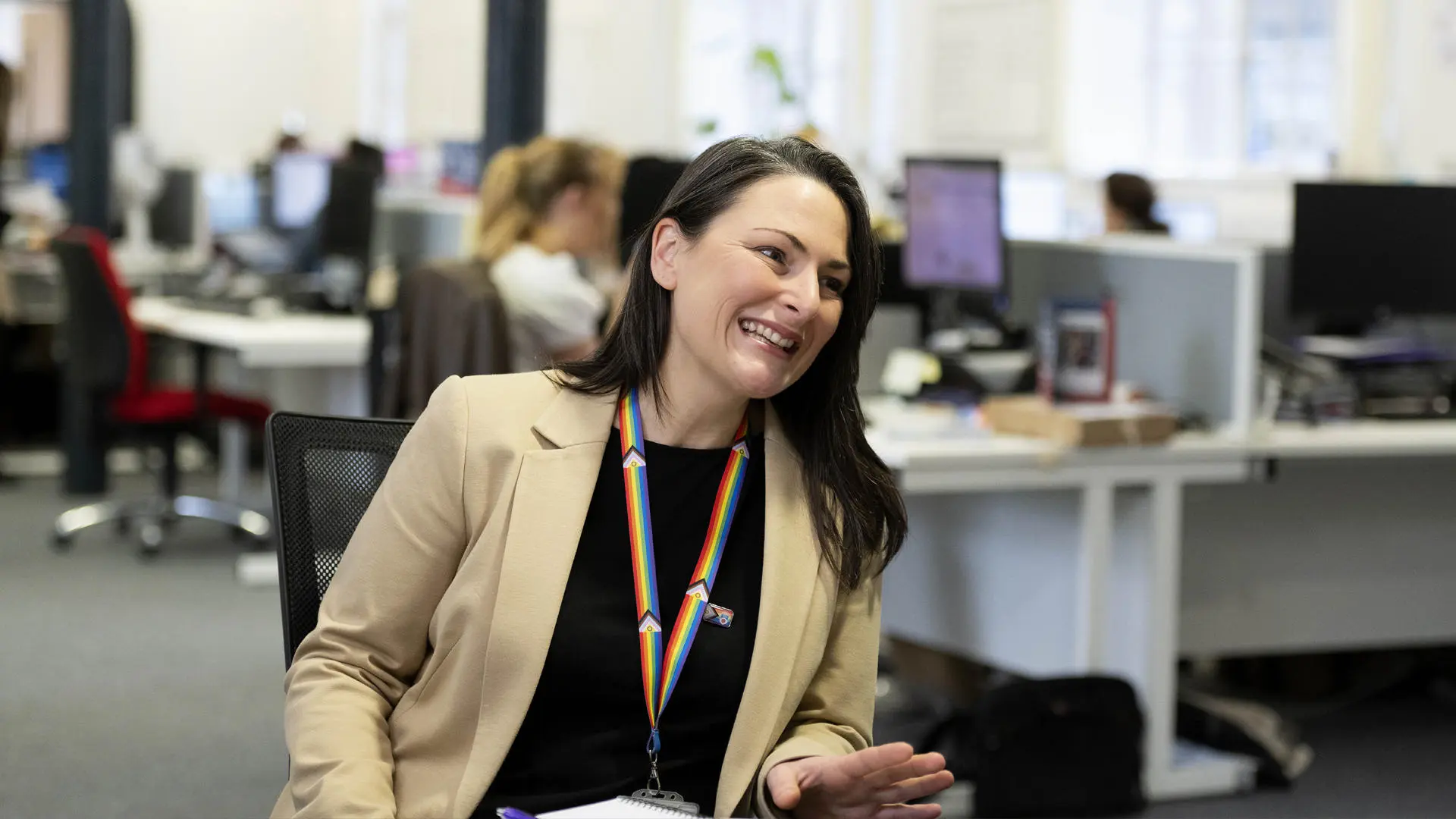 smiling staff member sat in an office building with computers and people in the background